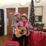 Two older women with pink cowboys hats play guitar in front of a Christmas Tree