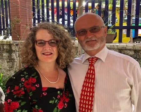 Anne Davies, a woman in her sixties, wearing a floral dress & glasses and standing next to Jeff, a man in his sixties wearing a white shirt and red & gold tie.