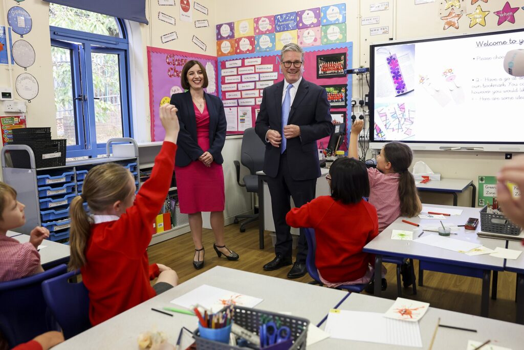 Two politicians stand in a classroom as children in the foreground raise their hands