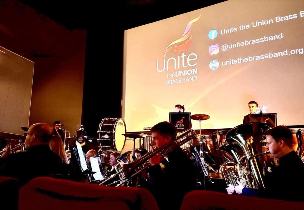 Brass band perform on a stage in front of a screen with the Unite the Union logo on it