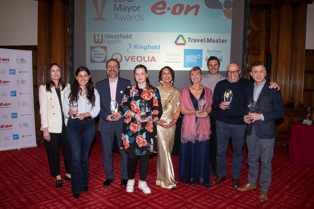 Nine people stand at an awards ceremony, six of them holding up their trophies