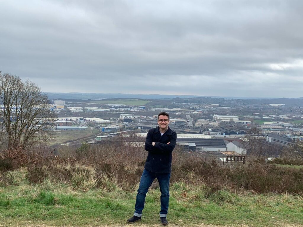 A man with glasses stands on a hill with a city background behind him