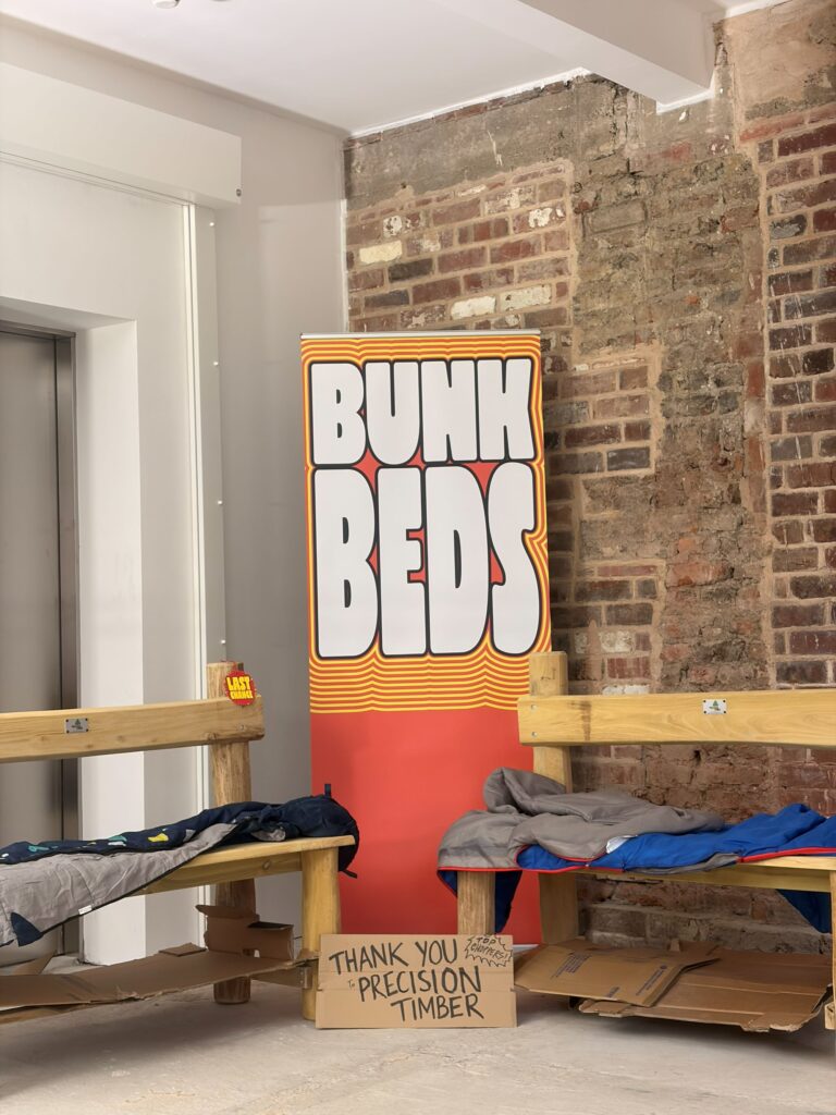 An image of wooden benches, with sleeping bags on them. In between them there is a red sign with 'Bunk Beds' in large white letters.   