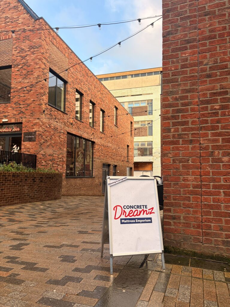 An image of a sign which reads 'Concrete Dreamz mattress emporium'  next to a brick wall. 