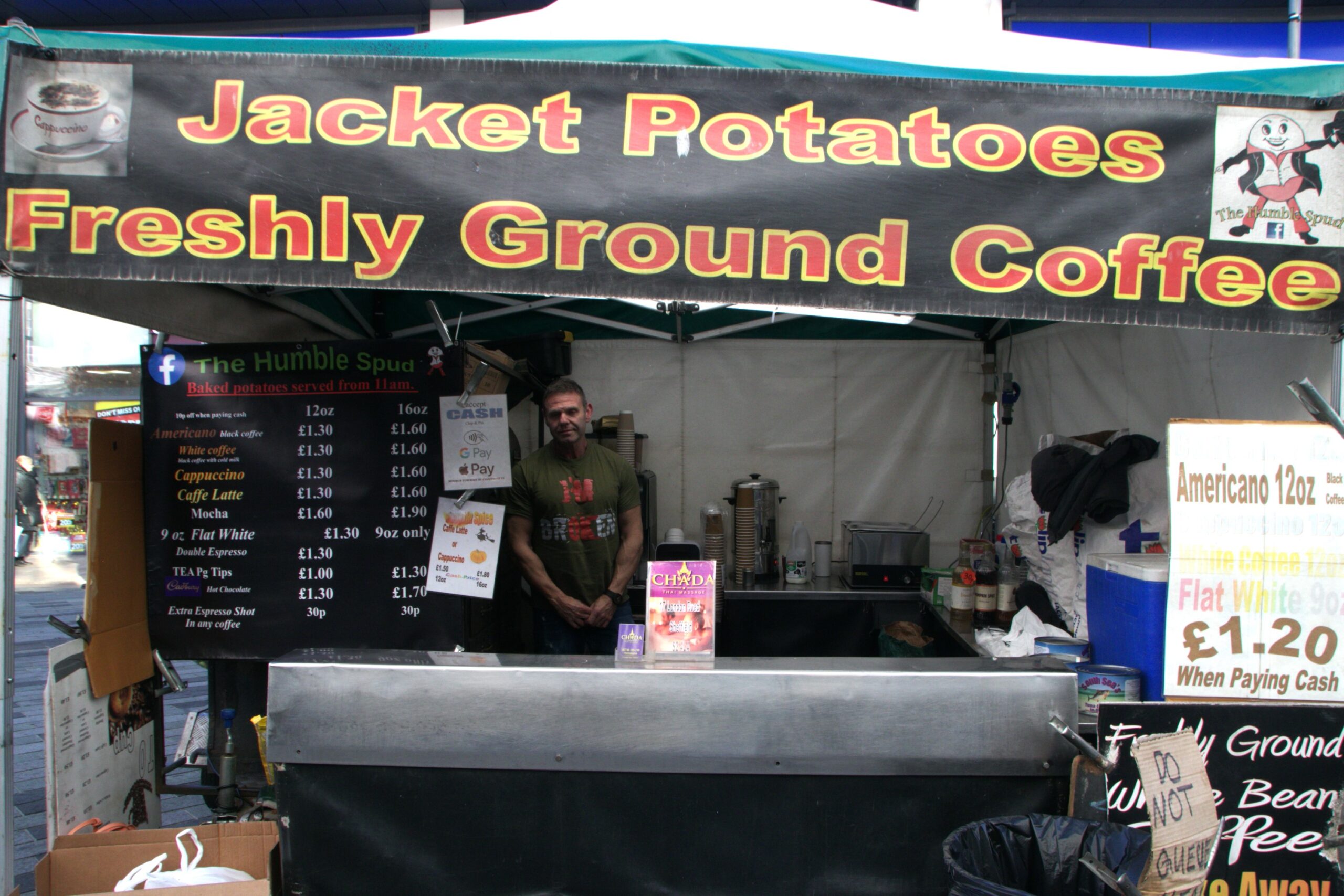 Man standing in jacket potato market stall with posters displaying list of jacket potatoes and hot drinks on offer