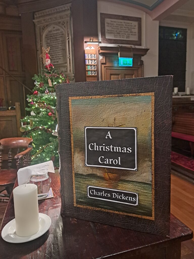 A large book which reads "A Christmas Carol Charles Dickens" set upon a dark brown wooden table next to a candle stick and a glass of water. 
