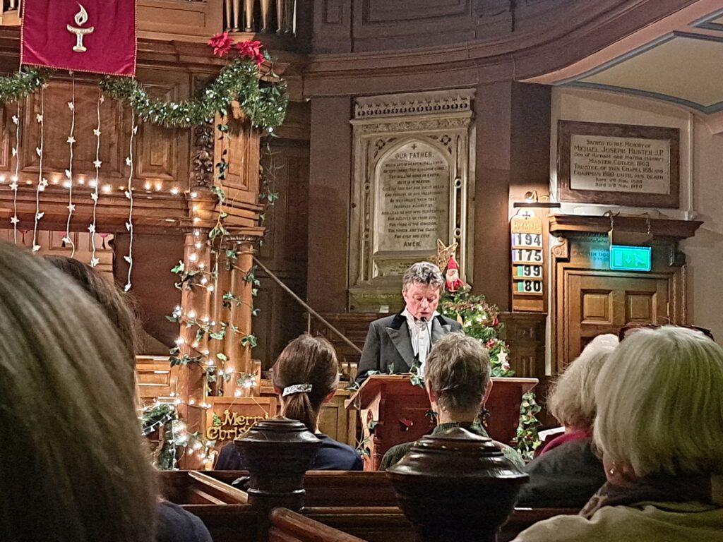 Patrick Smith reading aloud to the audience stood behind a pulpit. The church is decorated with fairy lights, tinsel and a Christmas tree.  