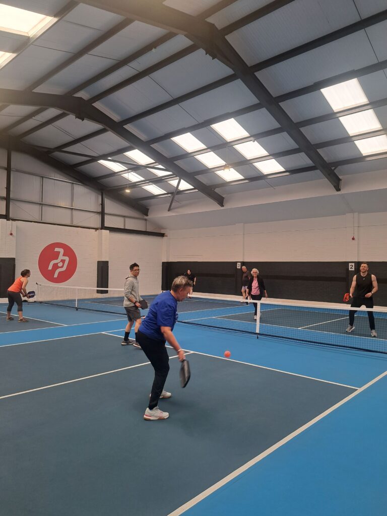 An inside pickleball blue court with seven players in shot holding rackets and hitting a plastic ball over a net 